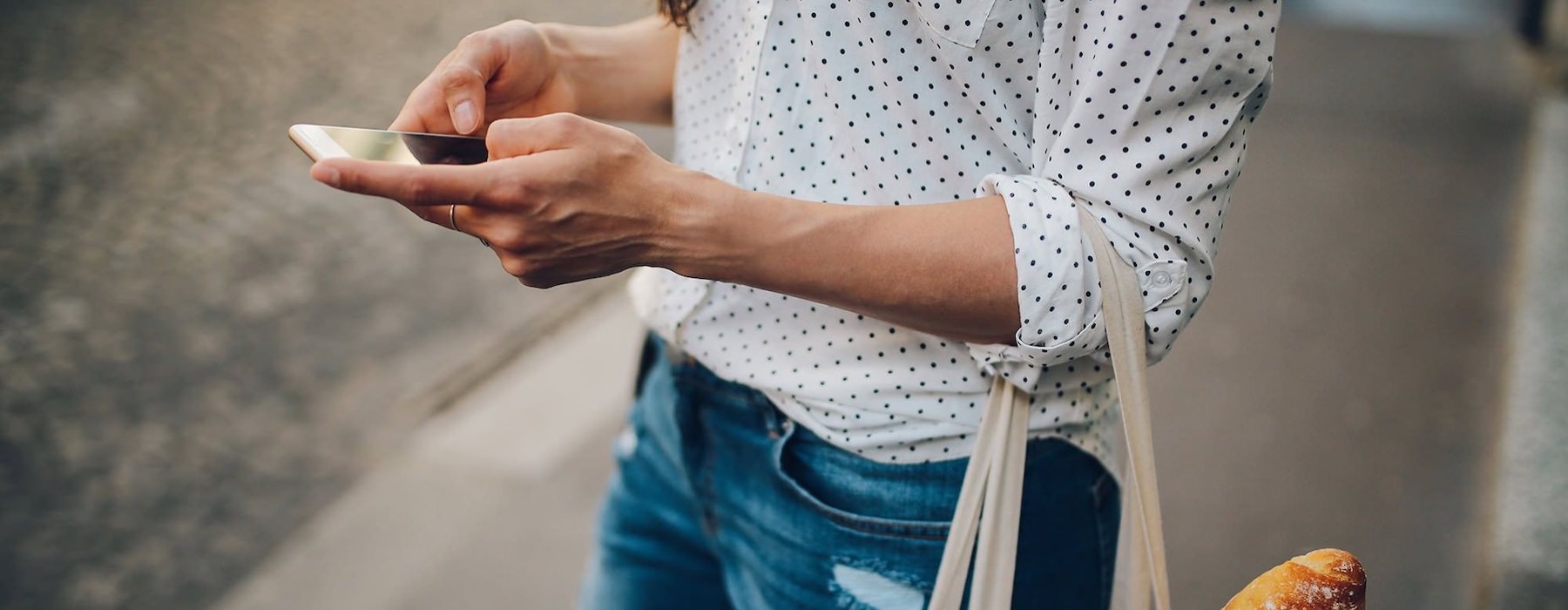 woman walks down the street and texts with a bag of groceries on her arm